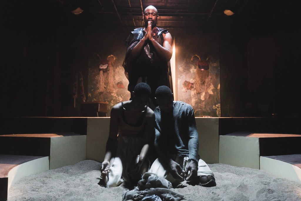 Three performers in a sand-filled ritual space, one standing with hands pressed together